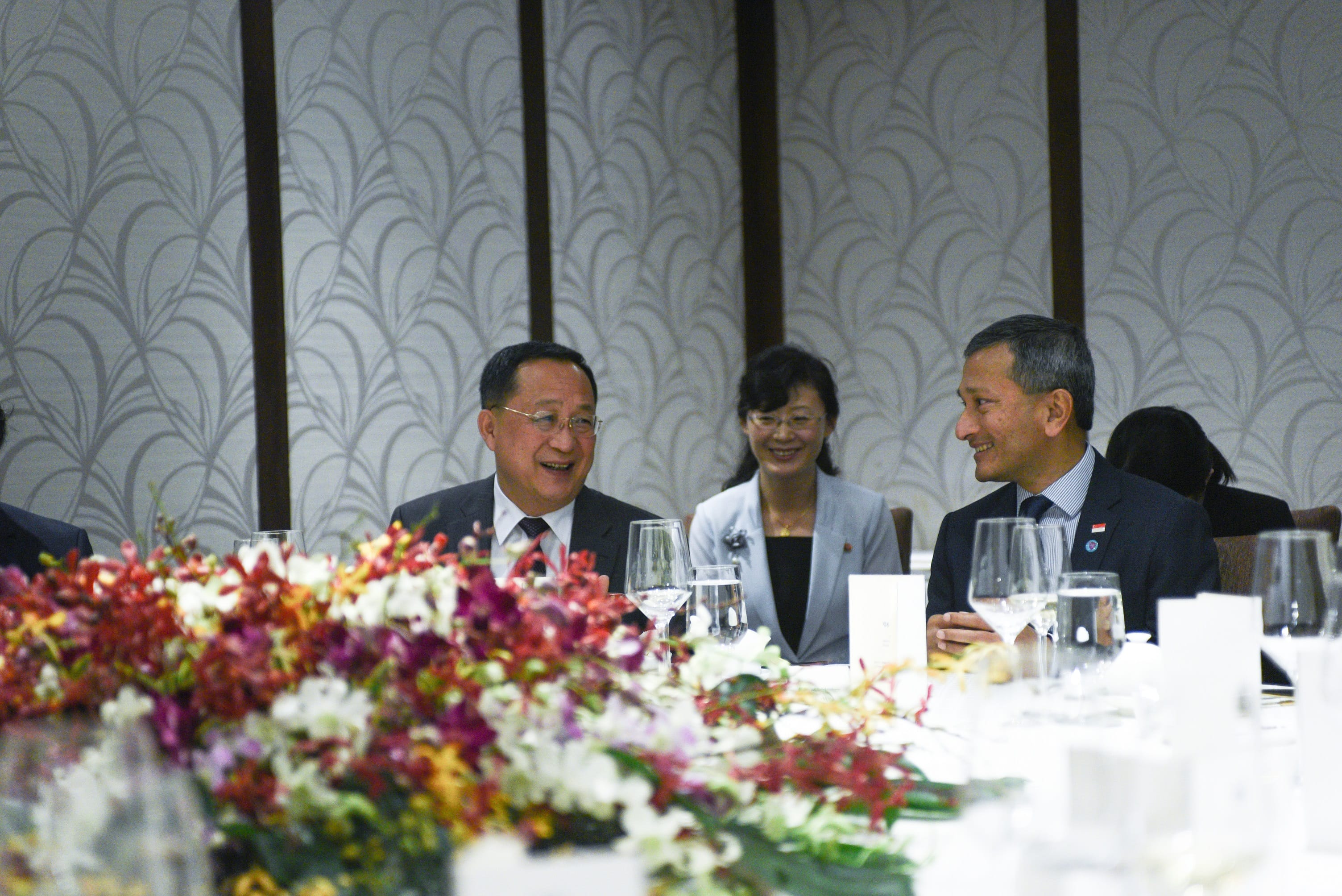 Two men in suits and a woman in a blazer sit at a table decorated with colorful flowers.